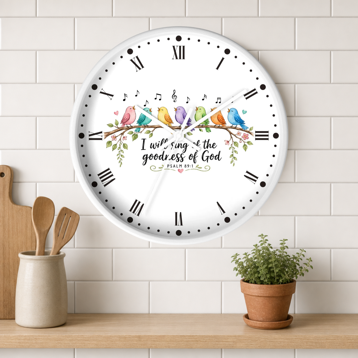 A round white wall clock with black Roman numerals and a colorful bird illustration across the center, mounted on a white subway tile wall above a wooden shelf with a small potted plant and utensils.