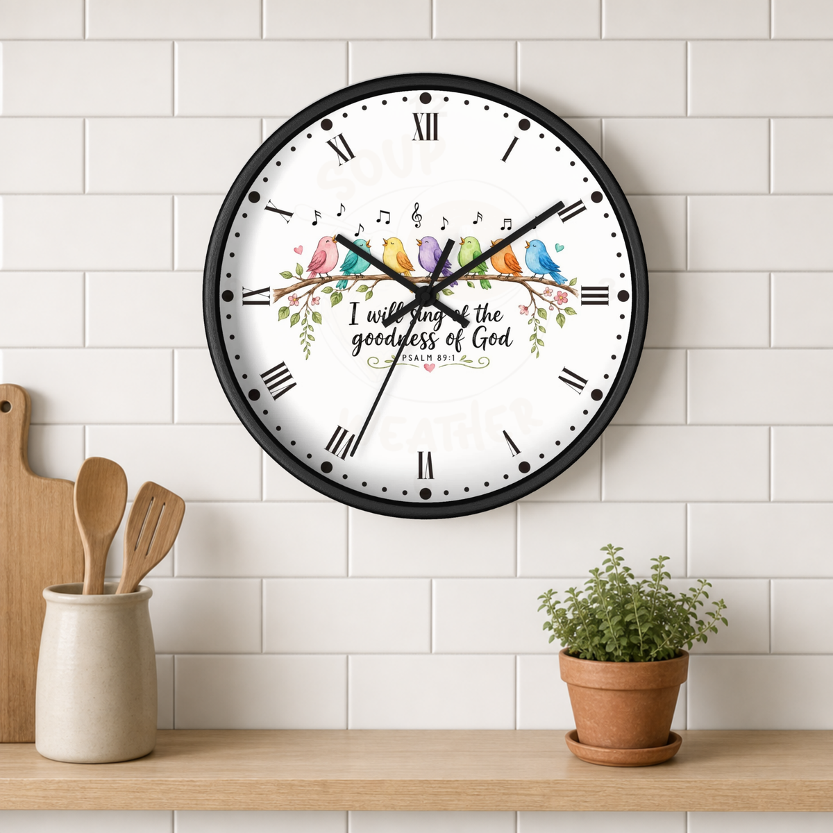 A round wall clock with a black frame and a white face, mounted on a white subway tile wall above a wooden shelf. The clock face features a colorful illustration of birds, musical notes, floral elements, and the text 'I will sing of the goodness of God' with 'PSALM 34:5' below.