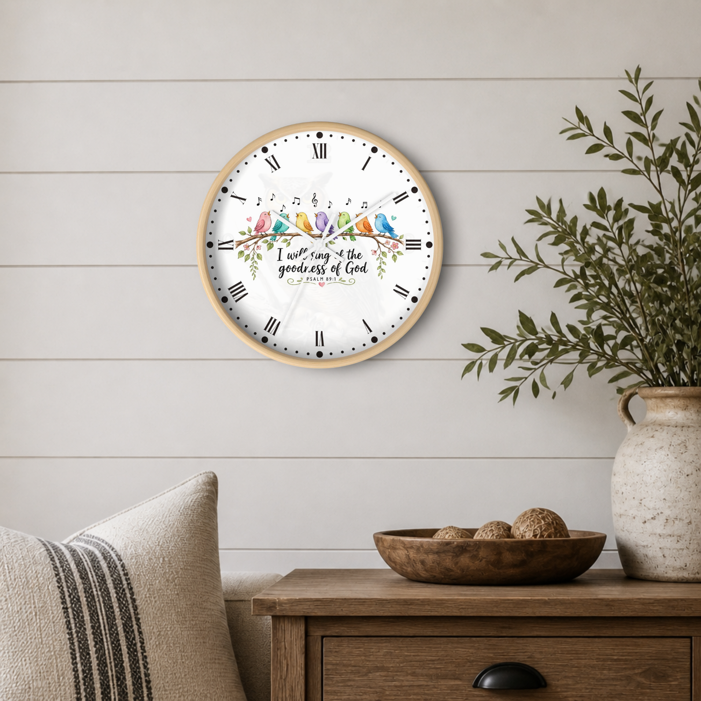 A round wall clock with a light wood frame and a white face hangs on a white shiplap wall above a wooden side table. The clock features a colorful illustration of birds perched on a branch with the text 'I will sing & the goodness of God is everlasting' and 'PSALM 100' in the center.