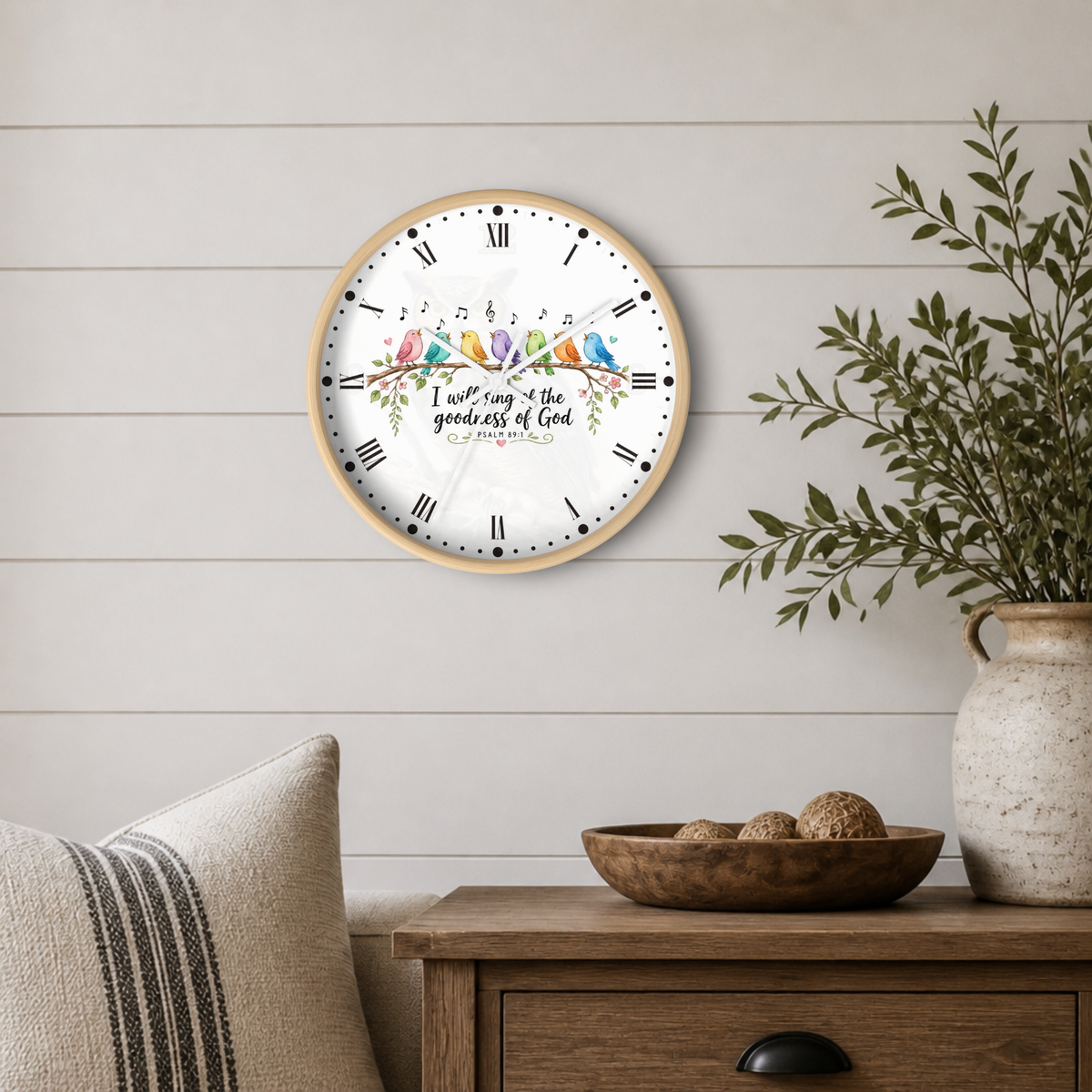 A round wall clock with a light wood frame and a white face hangs on a white shiplap wall above a wooden side table. The clock features a colorful illustration of birds perched on a branch with the text 'I will sing & the goodness of God is everlasting' and 'PSALM 100' in the center.