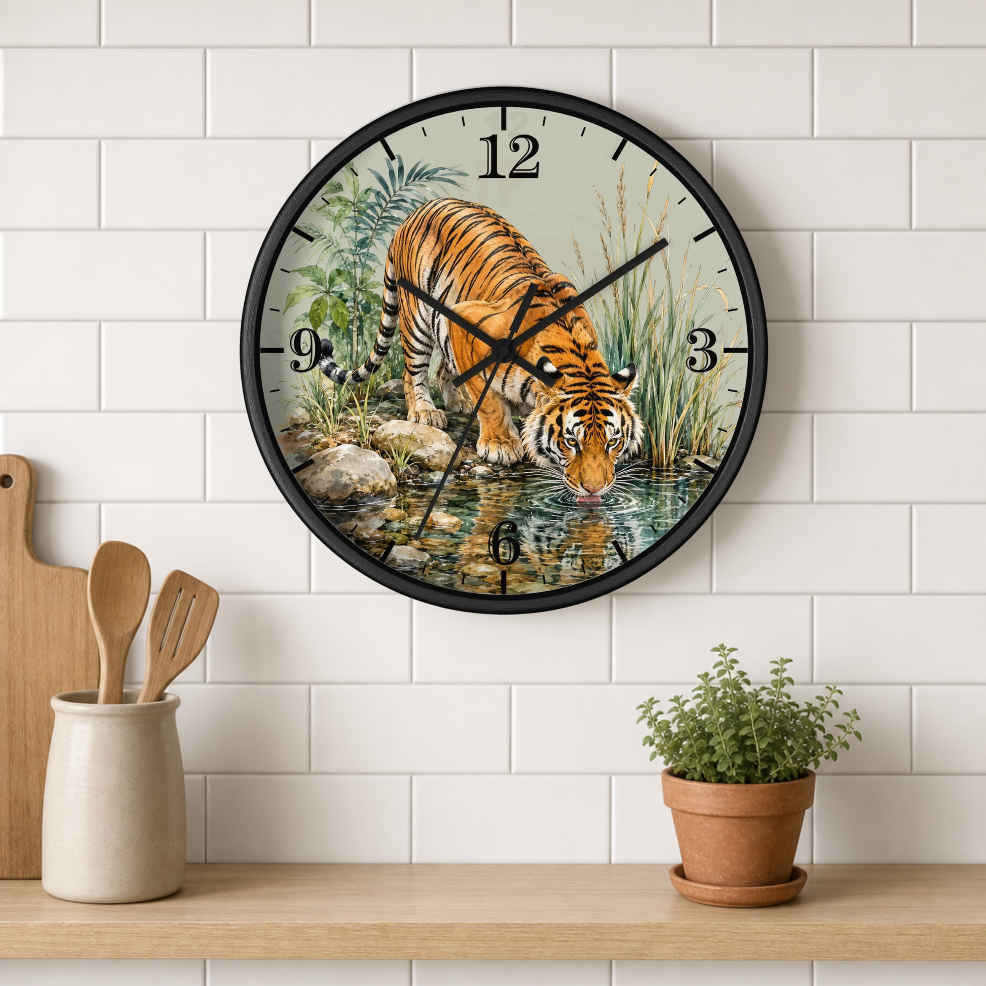 A round wall clock with a black frame hangs on a white subway-tile wall above a wooden shelf. The clock face features a detailed illustration of a tiger drinking water from a stream, surrounded by foliage and rocks.