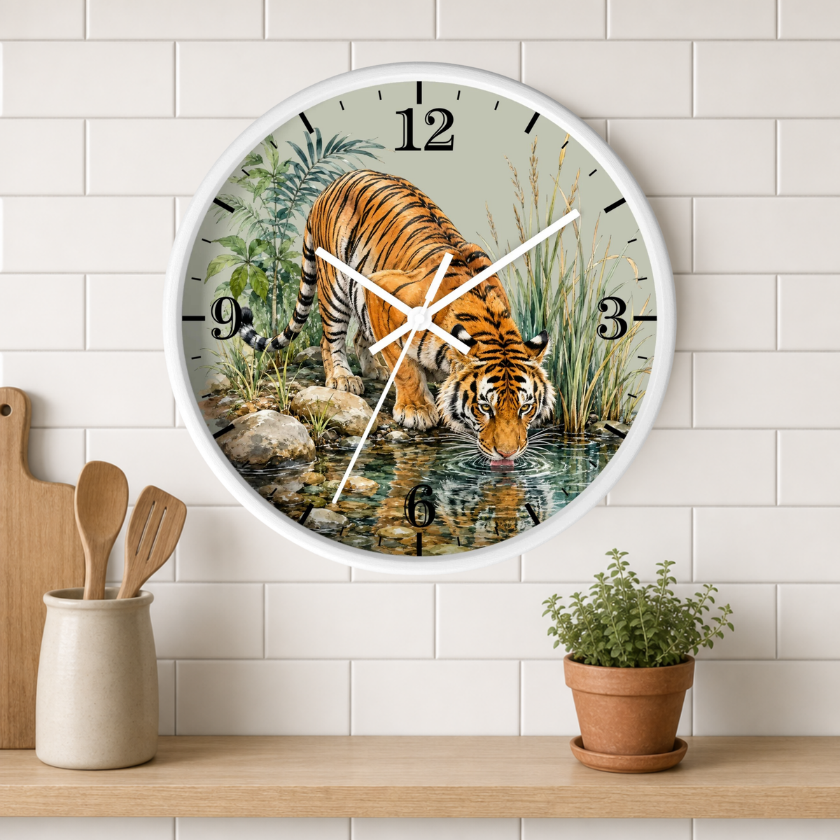 A round wall clock with a white frame and a printed illustration of a tiger drinking water, mounted on a white subway tile wall above a wooden shelf with a small potted plant and kitchen utensils.