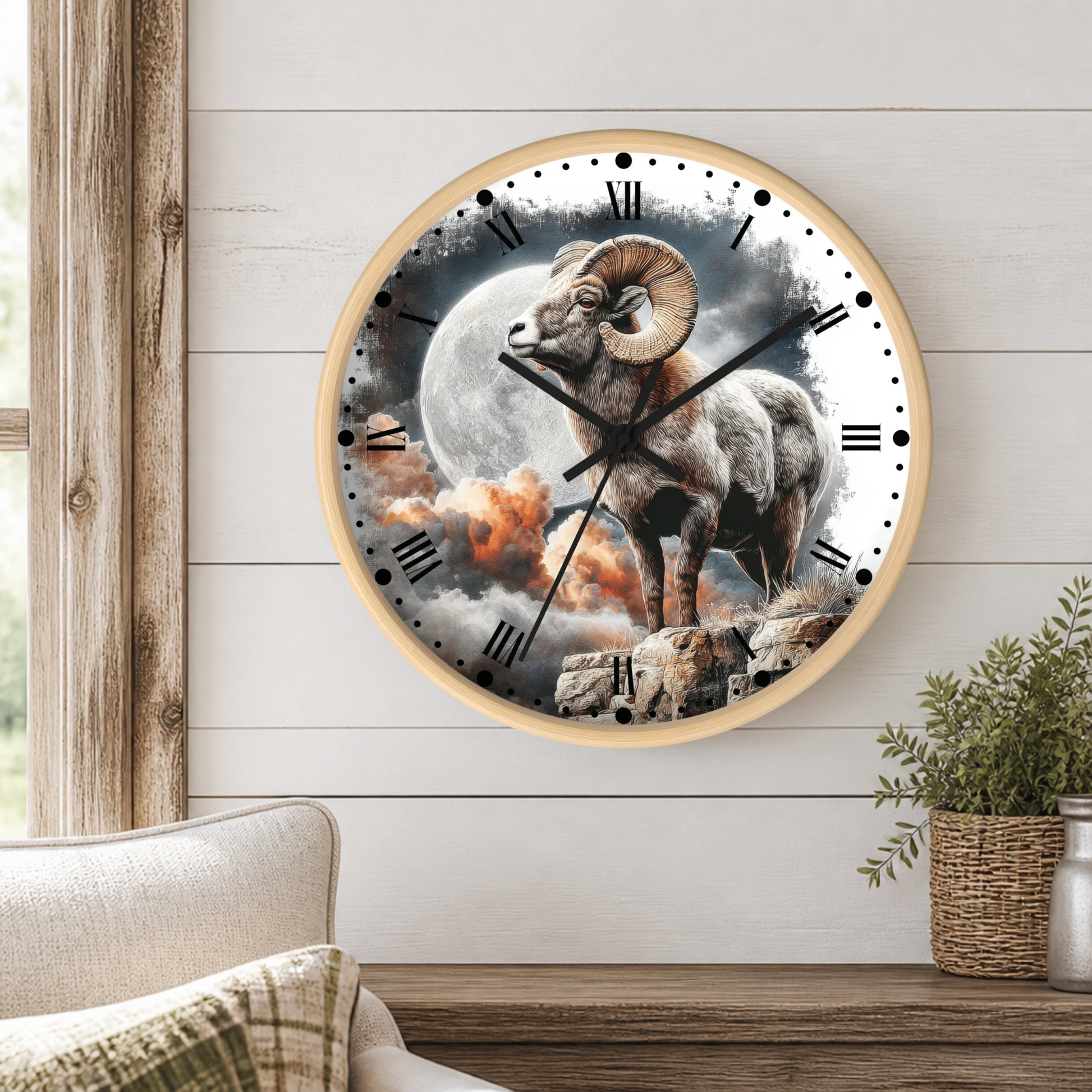 A round wall clock with a light wood frame hangs on a white shiplap wall above a wooden table. The clock face features a detailed graphic of a ram standing in front of a large full moon, with dark clouds and orange sun rays in the background.