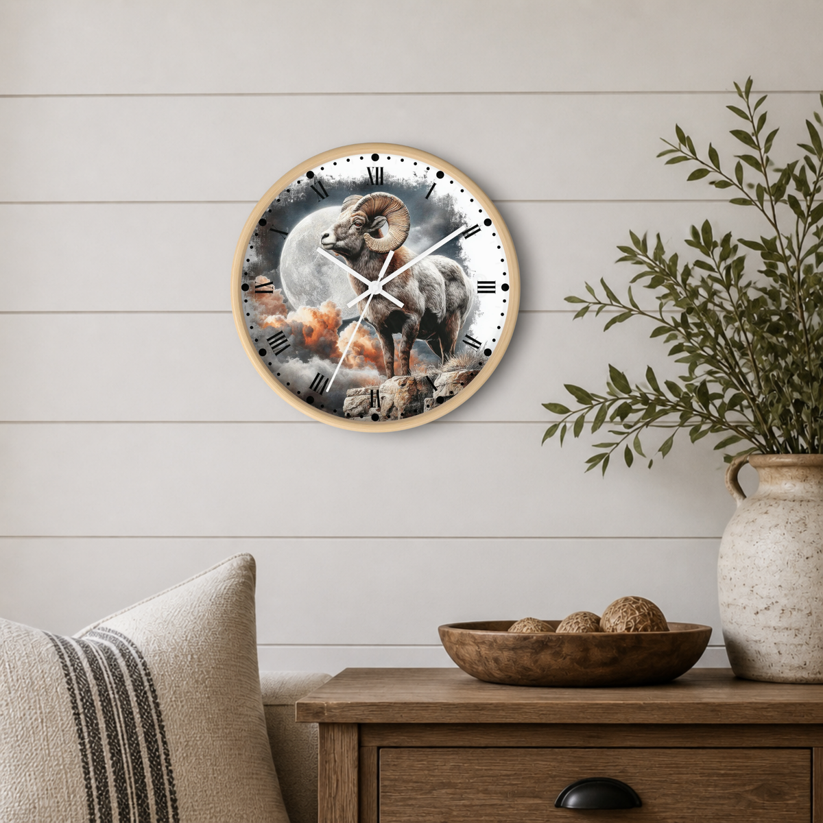 A round wall clock with a wood frame hangs on a white shiplap wall above a wooden side table. The clock face features a detailed illustration of a bighorn sheep standing in front of a full moon, with a small fire or embers near its feet.