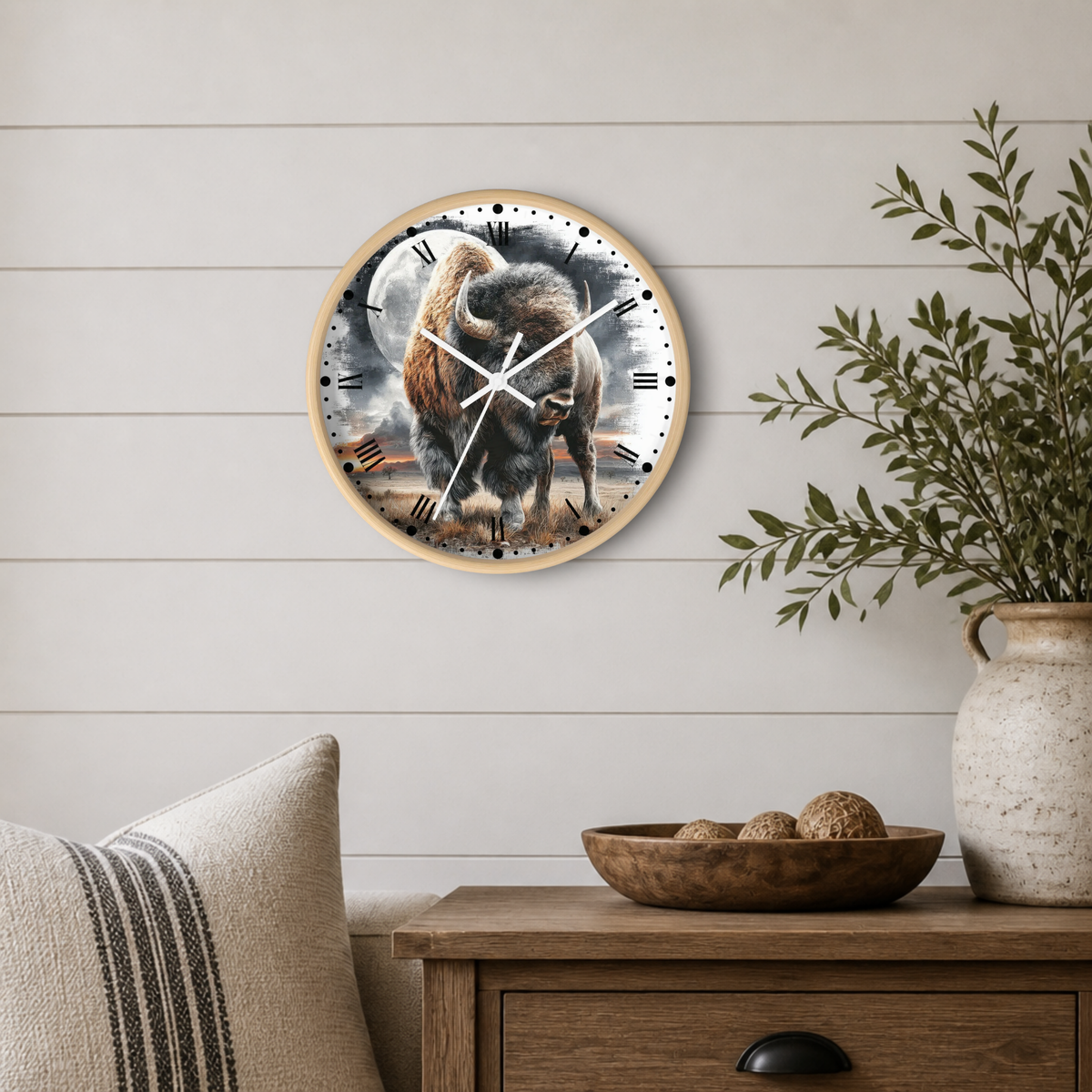 Round wall clock with a natural wood frame and a detailed bison illustration on the face, mounted on a white shiplap wall above a wooden side table with a plant and decorative bowl.
