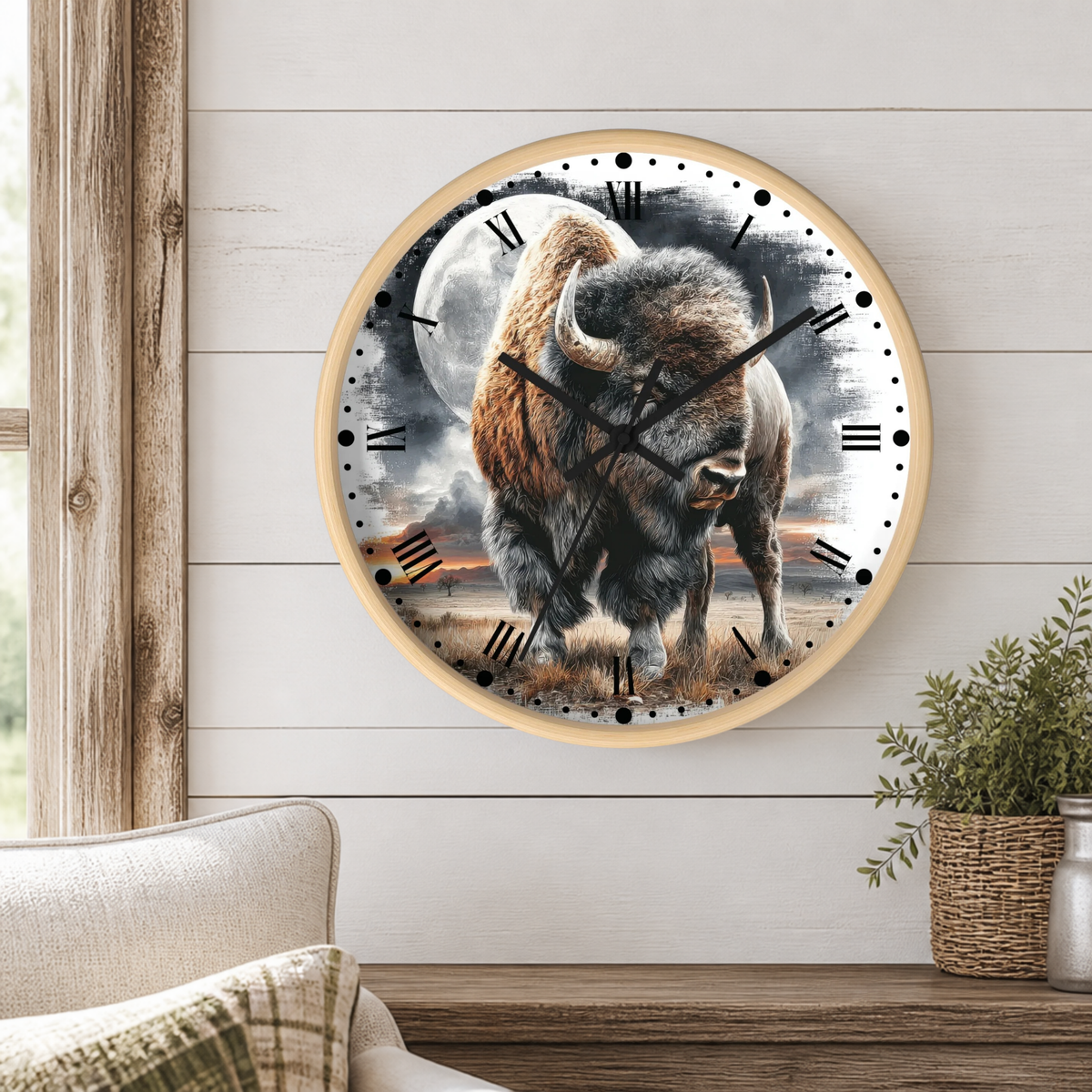 A round wall clock with a natural wood frame and a printed design of a bison walking in front of a full moon, mounted on a white subway tile wall above a wooden shelf with kitchen utensils and a potted plant.
