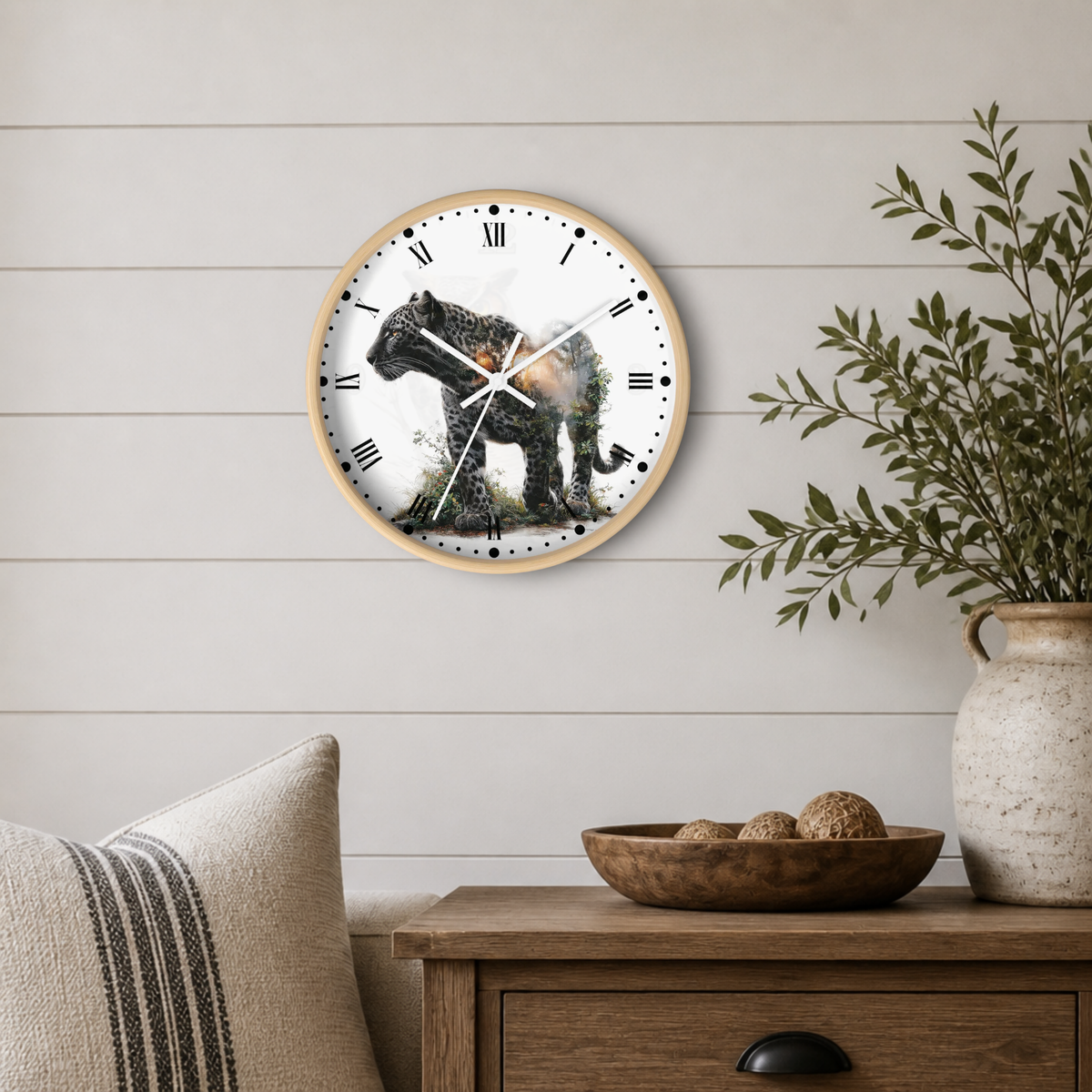 A round wall clock with a light wood frame and a white face featuring a black-and-white leopard illustration, mounted on a white shiplap wall above a wooden side table with a plant and bowl.