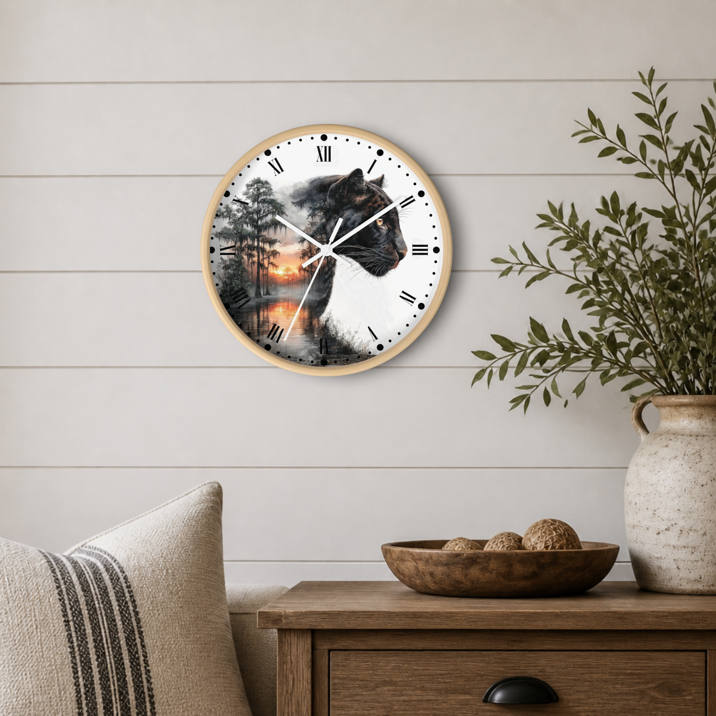 Round wall clock with a light wood frame and a printed design of a black panther over a forest and sunset scene, mounted on a white shiplap wall above a wooden side table with a plant and decorative bowl.