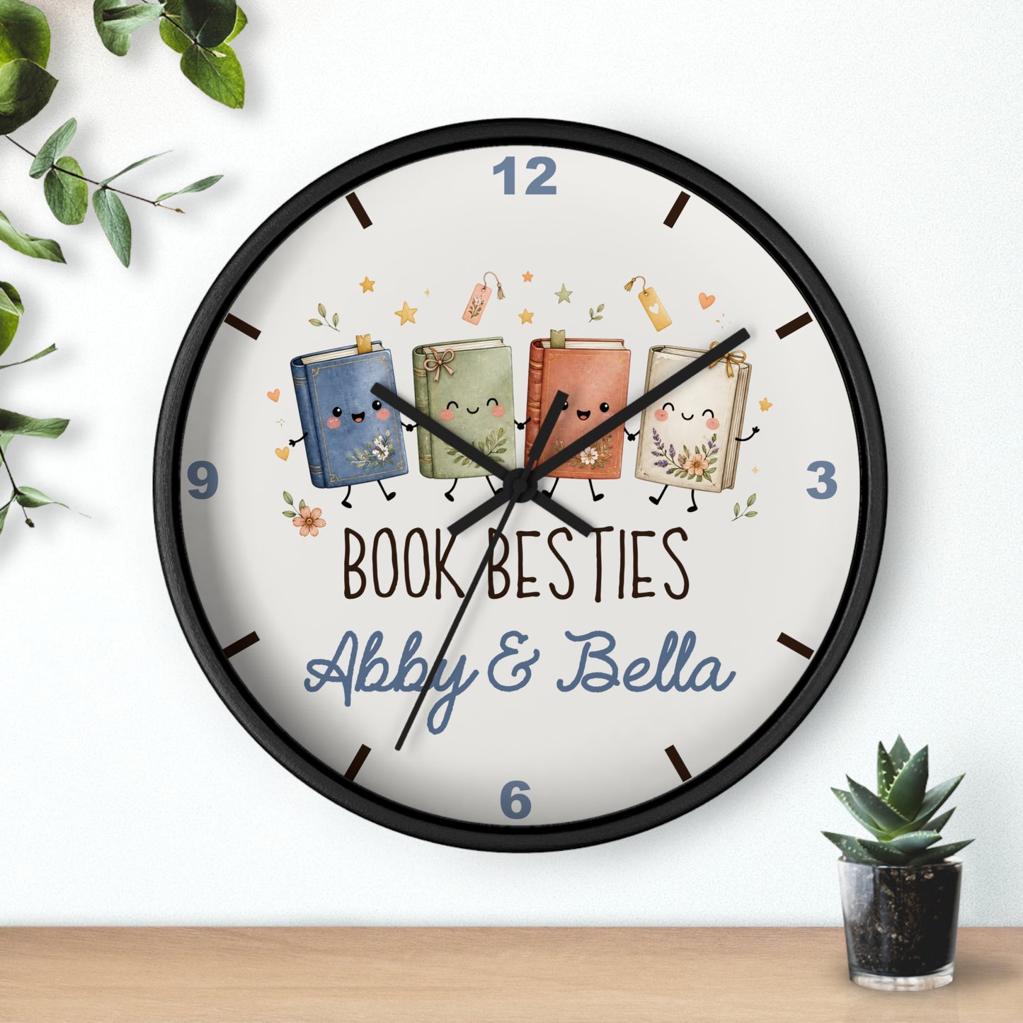 A round wall clock with a black frame and a white face, featuring four cute illustrated books with faces and the text 'BOOK BESTIES Abby & Bella'. The clock is shown hanging on a white wall above a wooden surface, with small green plants in the corners.
