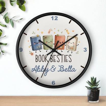 A round wall clock with a black frame and a white face, featuring four cute illustrated books with faces and the text 'BOOK BESTIES Abby & Bella'. The clock is shown hanging on a white wall above a wooden surface, with small green plants in the corners.