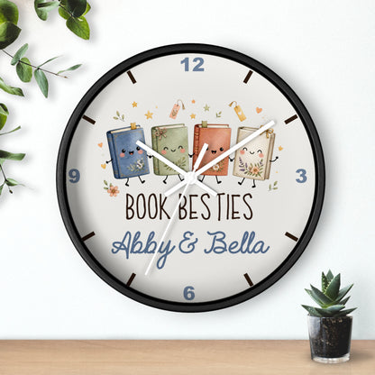 A round wall clock with a black frame and a white face, featuring three cute illustrated books with faces and the text 'BOOK BESTIES Abby & Bella'. The clock is shown hanging on a white wall above a wooden surface, with decorative green leaves and a small potted succulent nearby.