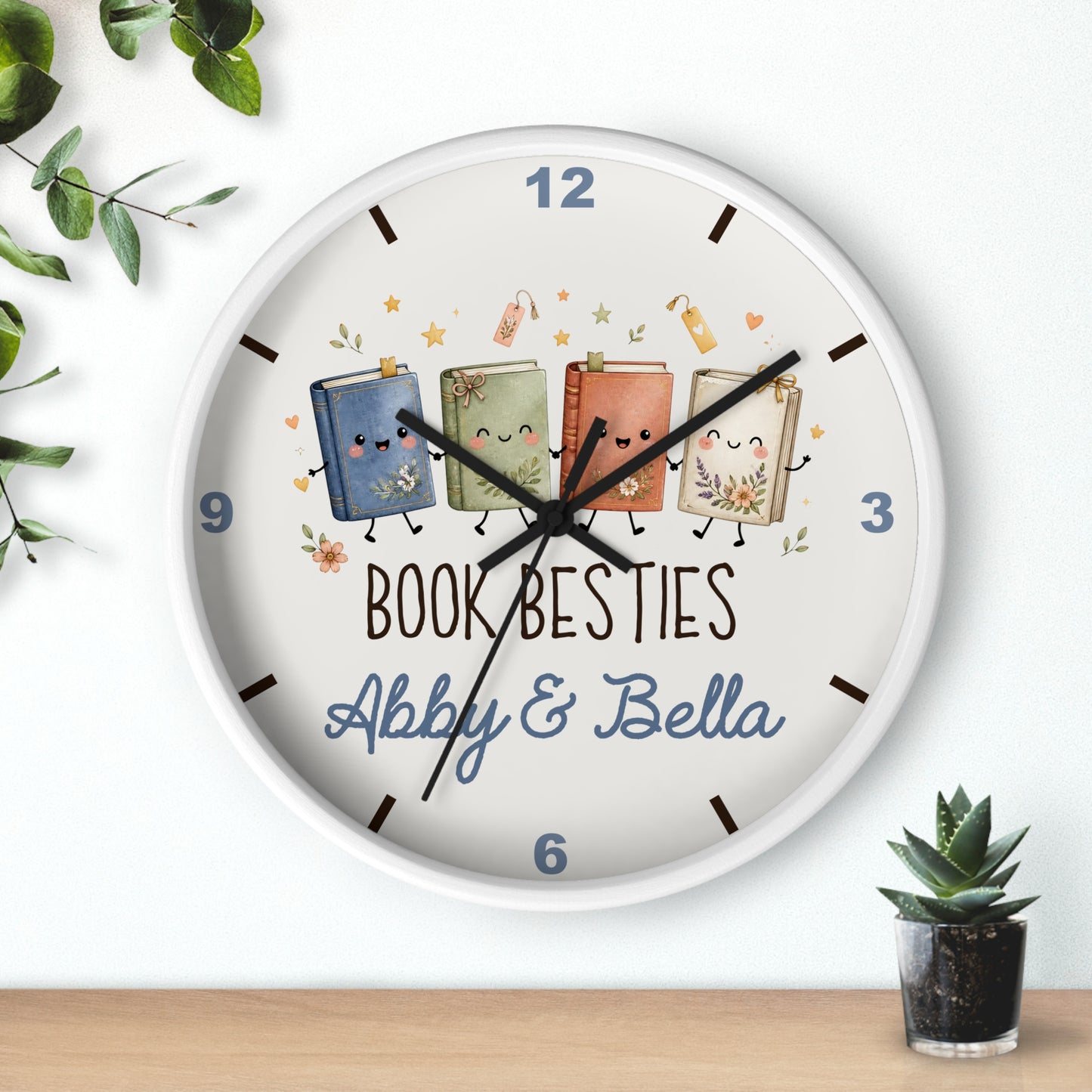 A round white wall clock with a playful book-themed design. The clock face features four cute, smiling cartoon books in blue, green, orange, and beige, with the text 'BOOK BESTIES Abby & Bella' printed below.
