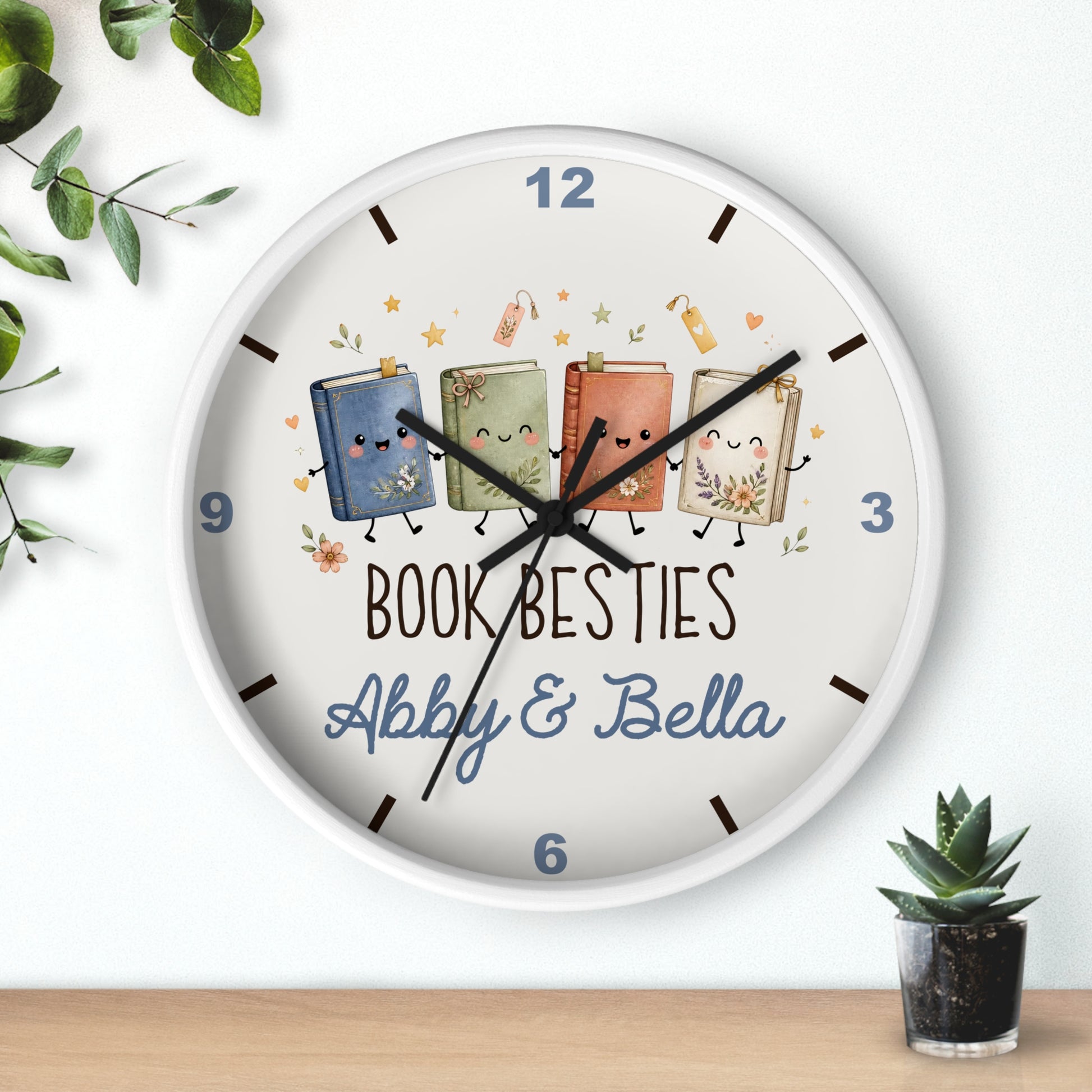 A round white wall clock with a playful book-themed design. The clock face features four cute, smiling cartoon books in blue, green, orange, and beige, with the text 'BOOK BESTIES Abby & Bella' printed below.