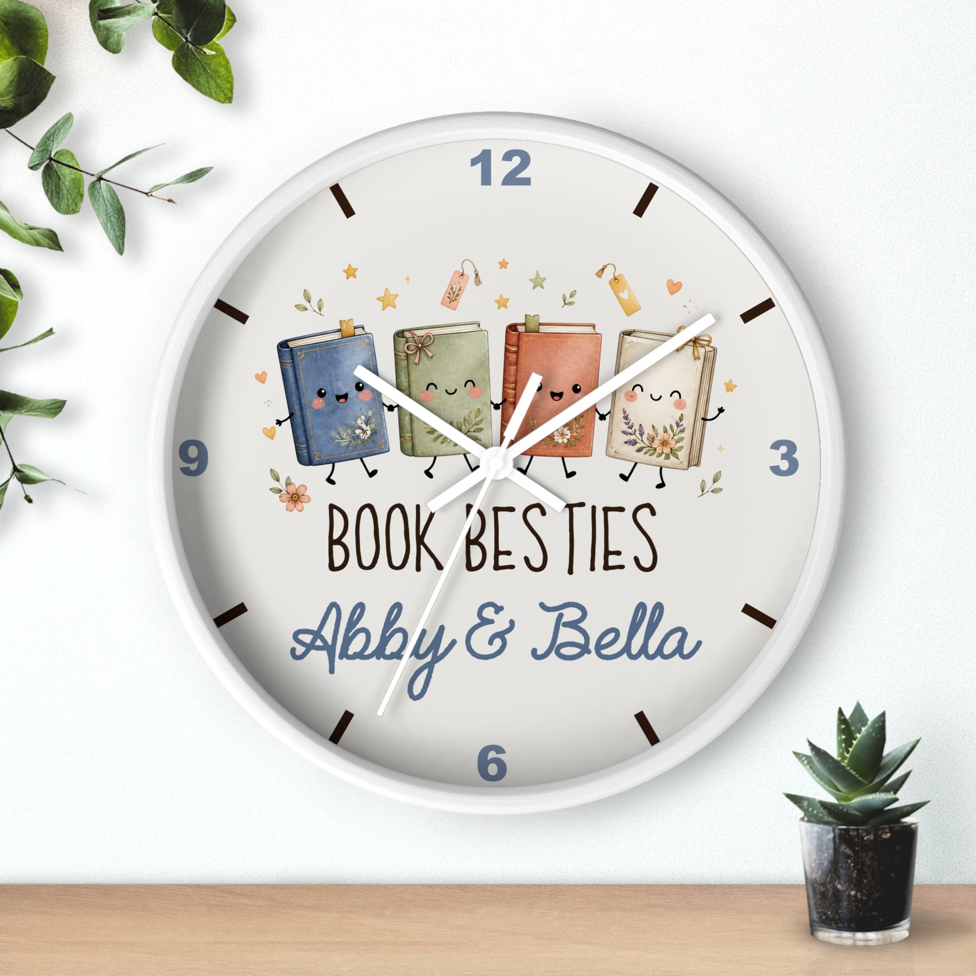 A round white wall clock with a whimsical illustration of four smiling cartoon books and the text 'BOOK BESTIES Abby & Bella' on the front. The clock is shown hanging on a white wall above a wooden surface, with decorative green leaves and a small potted succulent nearby.