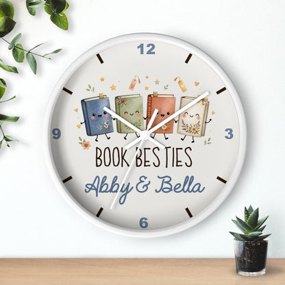 A round white wall clock with a whimsical illustration of four smiling cartoon books and the text 'BOOK BESTIES Abby & Bella' on the front. The clock is shown hanging on a white wall above a wooden surface, with decorative green leaves and a small potted succulent nearby.