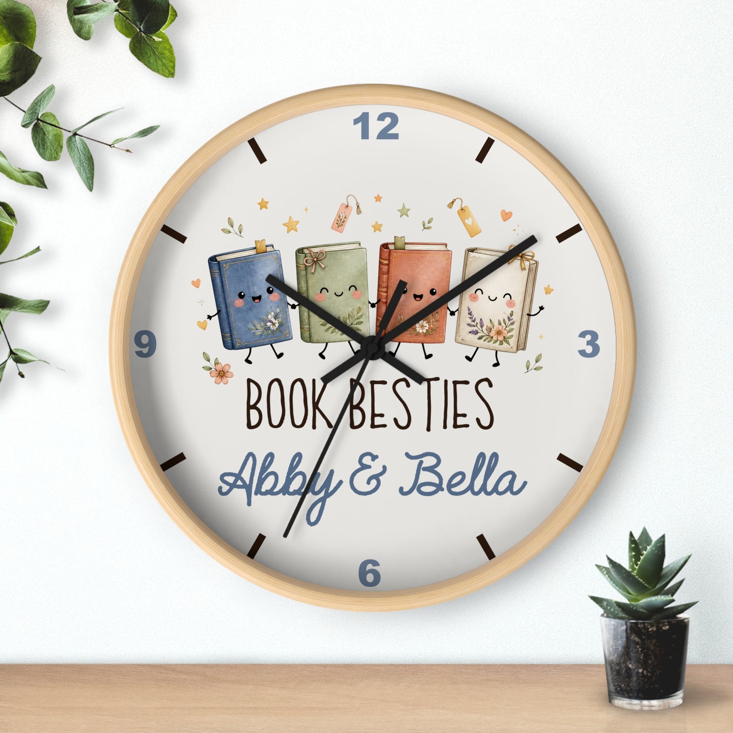 A round wooden wall clock with a white face, featuring a custom illustration of four cute, smiling books with faces and legs, along with the text 'BOOK BESTIES Abby & Bella'. The clock is shown hanging on a white wall above a wooden shelf, with decorative green leaves and a small potted succulent.