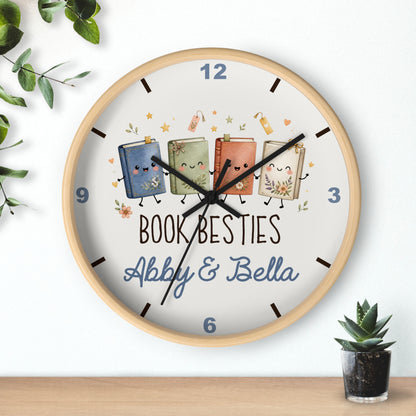 A round wooden wall clock with a white face, featuring a custom illustration of four cute, smiling books with faces and legs, along with the text 'BOOK BESTIES Abby & Bella'. The clock is shown hanging on a white wall above a wooden shelf, with decorative green leaves and a small potted succulent.