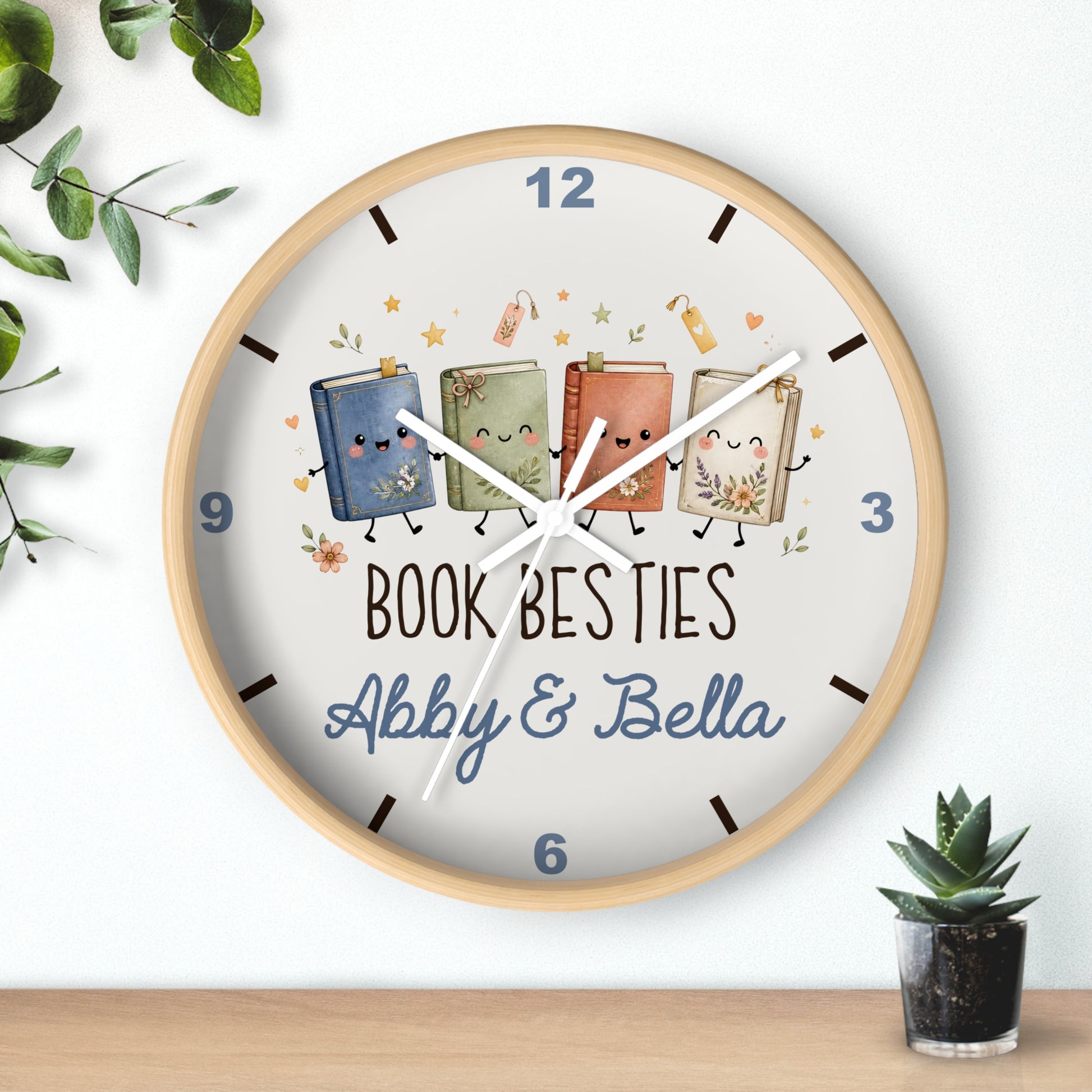 A round wall clock with a light wood frame and a white face, featuring three cute illustrated books with faces and the text 'BOOK BESTIES Abby & Bella'. The clock is shown hanging on a white wall above a wooden surface, with decorative green leaves and a small potted succulent nearby.
