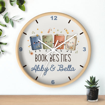 A round wall clock with a light wood frame and a white face, featuring three cute illustrated books with faces and the text 'BOOK BESTIES Abby & Bella'. The clock is shown hanging on a white wall above a wooden surface, with decorative green leaves and a small potted succulent nearby.
