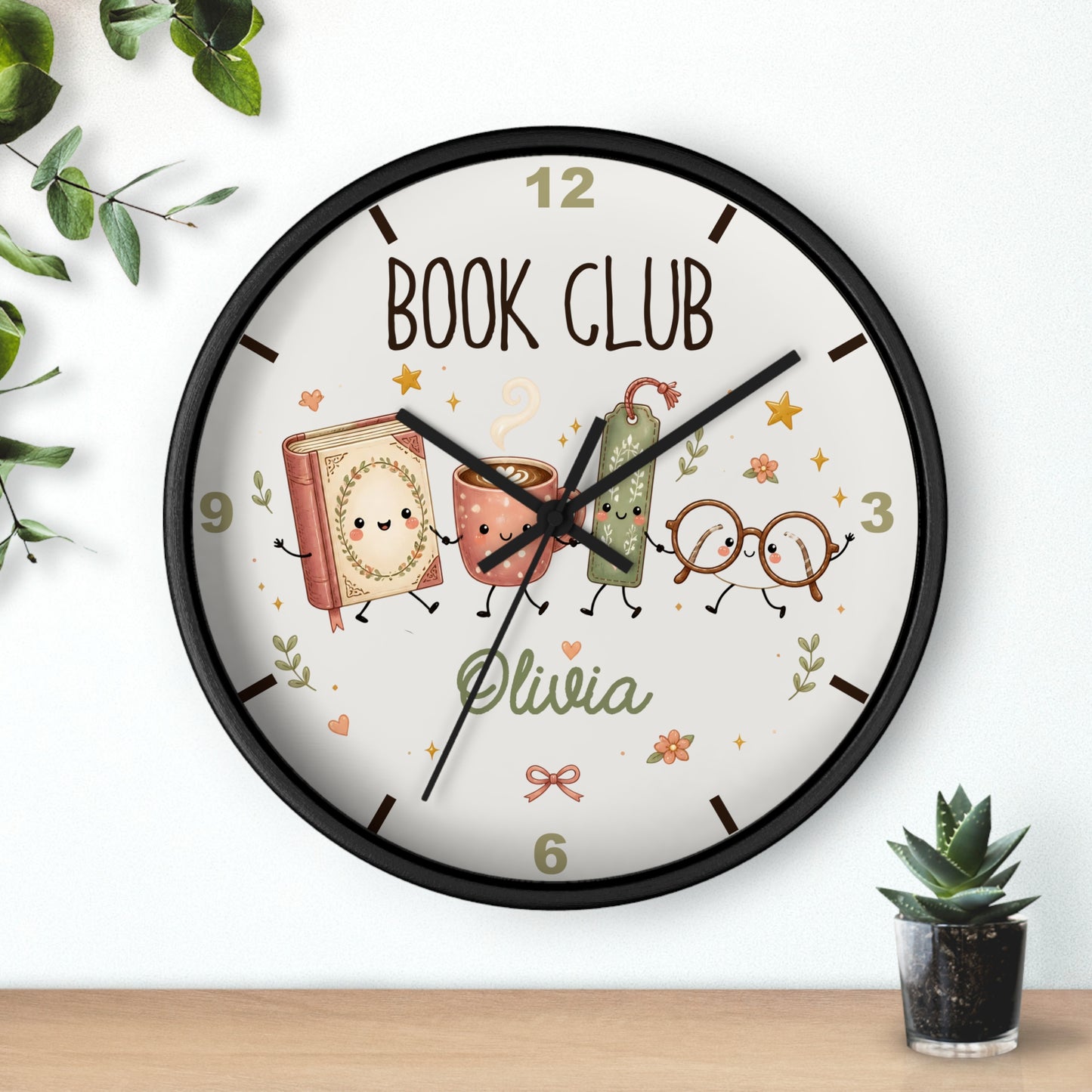 A round wall clock with a black frame and a white face featuring a personalized 'BOOK CLUB' design with cartoon books, glasses, and the name 'Olivia'. The clock is shown hanging on a white wall above a wooden shelf with small plants.