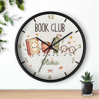 A round wall clock with a black frame and a white face featuring a personalized 'BOOK CLUB' design with cartoon books, glasses, and the name 'Olivia'. The clock is shown hanging on a white wall above a wooden shelf with small plants.