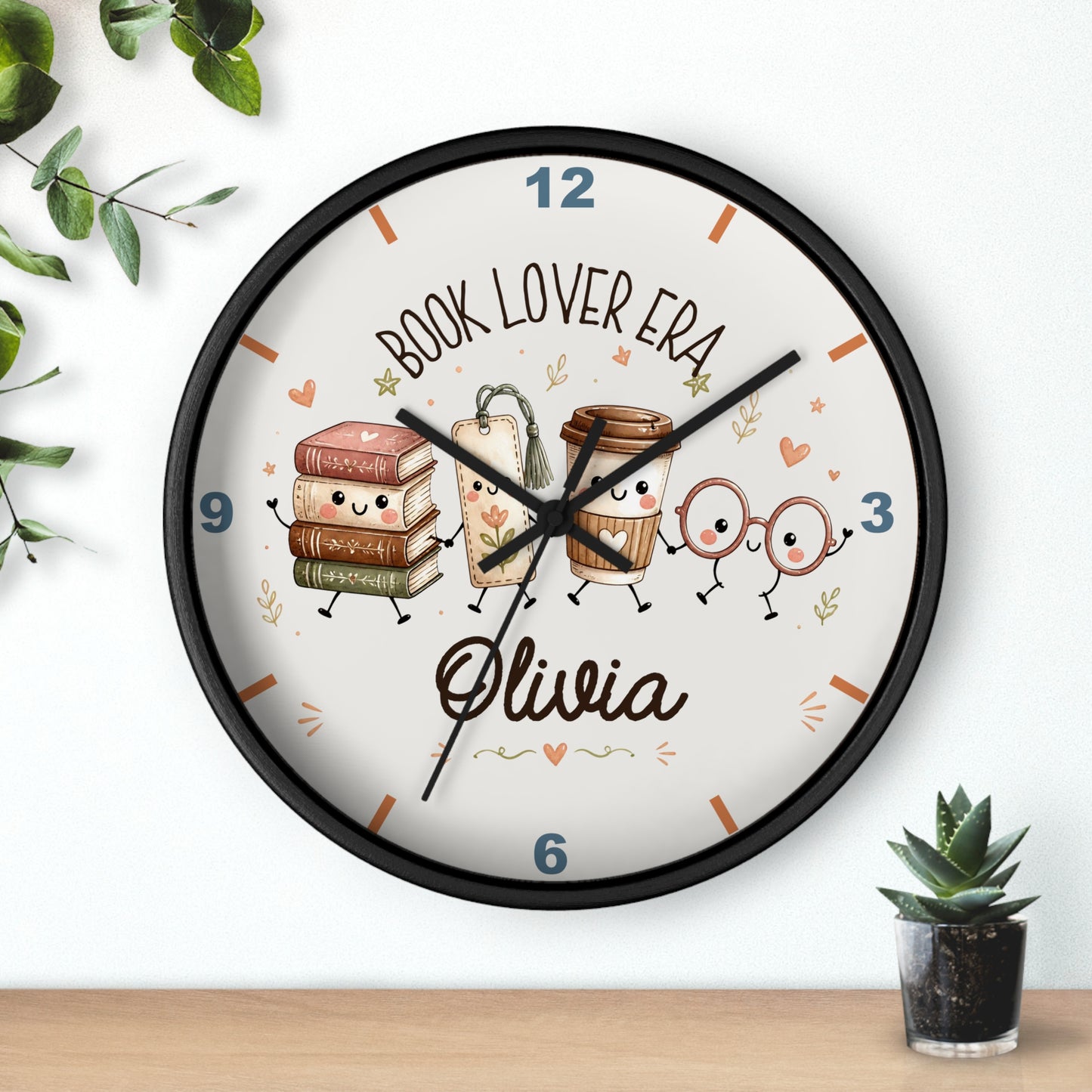 A round wall clock with a black frame and a white face, decorated with cute illustrations of books, coffee cups, and glasses on a shelf above a small potted succulent. The clock face features the text 'BOOK LOVER ERA' and the name 'Olivia' in script.