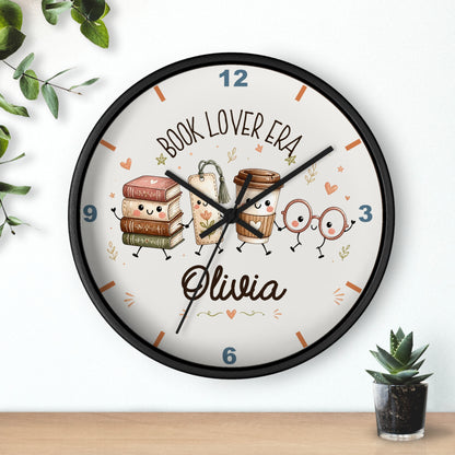 A round wall clock with a black frame and a white face, decorated with cute illustrations of books, coffee cups, and glasses on a shelf above a small potted succulent. The clock face features the text 'BOOK LOVER ERA' and the name 'Olivia' in script.