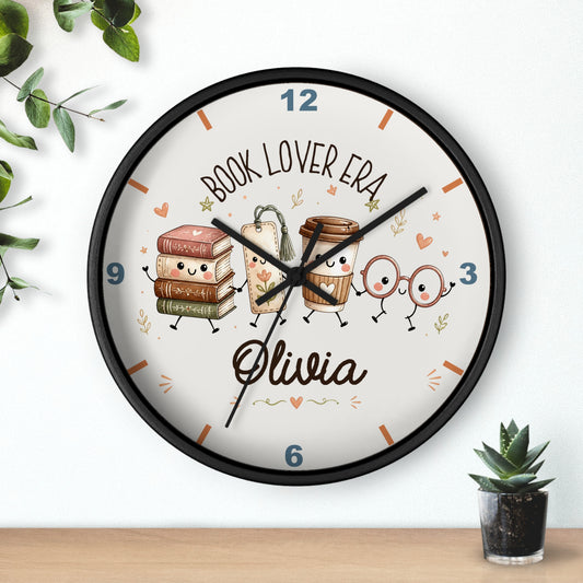 A round wall clock with a black frame and a white face, decorated with cute illustrations of books, coffee cups, and glasses on a shelf above a small potted succulent. The clock face features the text 'BOOK LOVER ERA' and the name 'Olivia' in script.