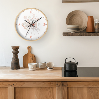 A round wall clock with a light wood frame and a white face featuring a delicate floral illustration and red numerals, styled in a warm kitchen setting above a wooden counter with ceramic dishes and a black kettle.