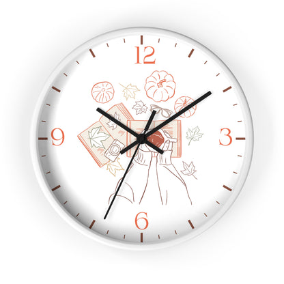 A round wall clock with a white face and outer rim, featuring black hands and orange numerals. The center of the dial has a delicate line-art illustration of a woman holding books with autumn leaves and pumpkins.