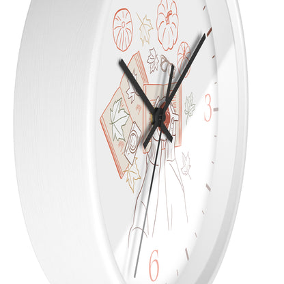 A close-up of a white round wall clock face with black hands and a minimalist autumn-themed illustration featuring orange numerals and leaf motifs.