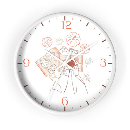 A round wall clock with a white face and silver-toned outer rim, featuring a minimalist line-art illustration of a person holding a red book surrounded by autumn leaves, pumpkins, and books in muted orange and brown tones. The numbers 1 through 9 and 6 through 12 are printed in orange, with brown minute markers and white hands.
