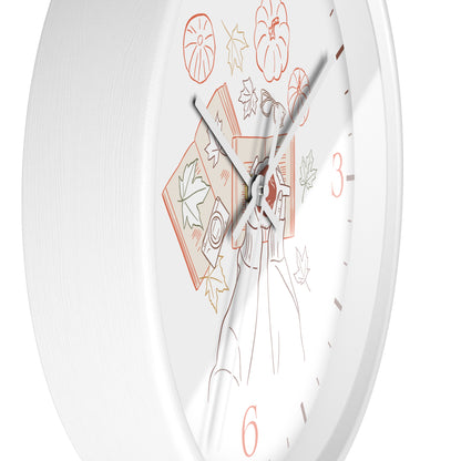 A close-up of a white round wall clock face with minimalist autumn-themed line art, including a pumpkin, leaves, and a woman reading, with coral-colored numerals and hands.