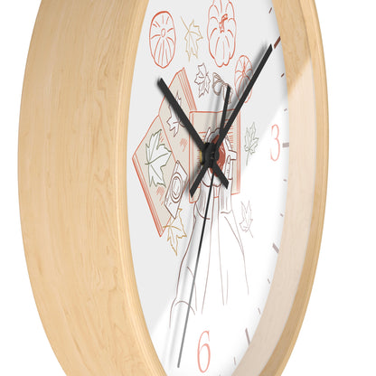 A close-up of a wall clock with a light natural wood frame and a white dial decorated with autumn-themed illustrations, including pumpkins, leaves, and a figure in a long coat. The clock has black hands and red numerals.