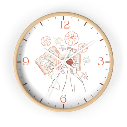 A round wall clock with a light natural wood frame and a white face featuring an autumn-themed illustration of a person holding a book, pumpkins, leaves, and orange numerals.