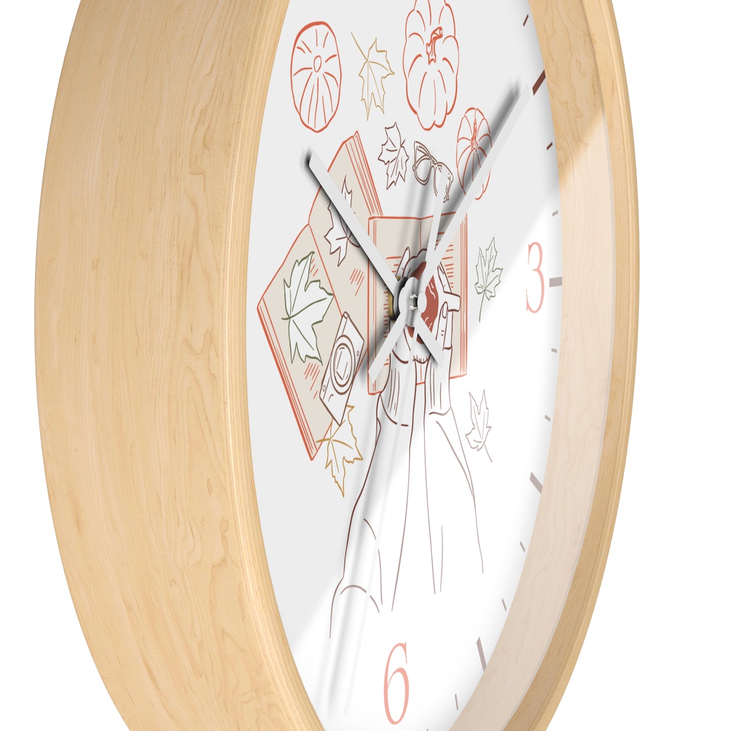 A close-up of a wall clock with a light natural wood frame and a white dial featuring autumn-themed illustrations, including pumpkins, leaves, and a witch's hat, with red numerals at 3 and 6.