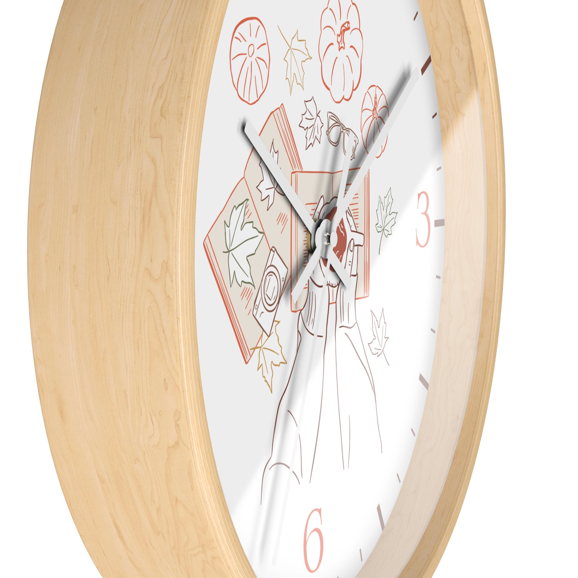 A close-up of a wall clock with a light natural wood frame and a white dial featuring autumn-themed illustrations, including pumpkins, leaves, and a witch's hat, with red numerals at 3 and 6.