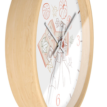 A close-up of a wall clock with a light natural wood frame and a white dial featuring autumn-themed illustrations, including pumpkins, leaves, and a witch's hat, with red numerals at 3 and 6.