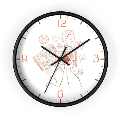 A round wall clock with a black frame and a white face featuring a minimalist line art illustration of a woman holding books and pumpkins, with orange numerals and black hands.