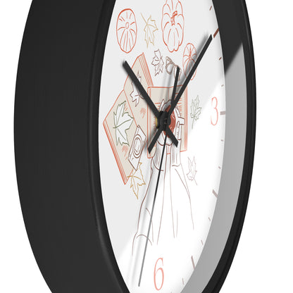 A close-up of a round wall clock with a black frame and a white face featuring autumn-themed line drawings, including pumpkins, leaves, and a figure, with black and red numerals.