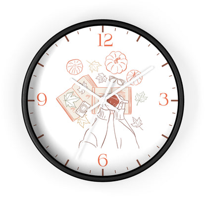 A round wall clock with a black frame and a white face featuring an autumn-themed illustration of a person holding a book, pumpkins, leaves, and orange numerals.