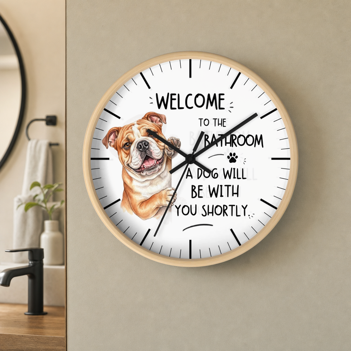 A round wall clock with a light wood frame hangs on a beige wall in a bathroom setting. The clock face is white with black text reading 'WELCOME TO THE BATHROOM A DOG WILL BE WITH YOU SHORTLY' and a painted bulldog illustration on the left side.