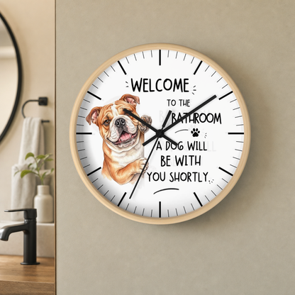 A round wall clock with a light wood frame hangs on a beige wall in a bathroom setting. The clock face is white with black text reading 'WELCOME TO THE BATHROOM A DOG WILL BE WITH YOU SHORTLY' and a painted bulldog illustration on the left side.