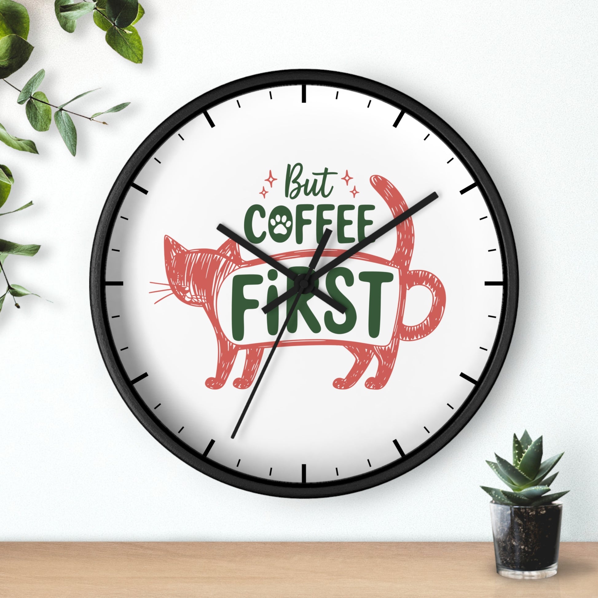 A round wall clock with a black frame and a white face, featuring the phrase 'But COFFEE FIRST' in green text with a red cat illustration. The clock is mounted on a white wall above a light wood surface, with decorative green leaves and a small potted succulent nearby.