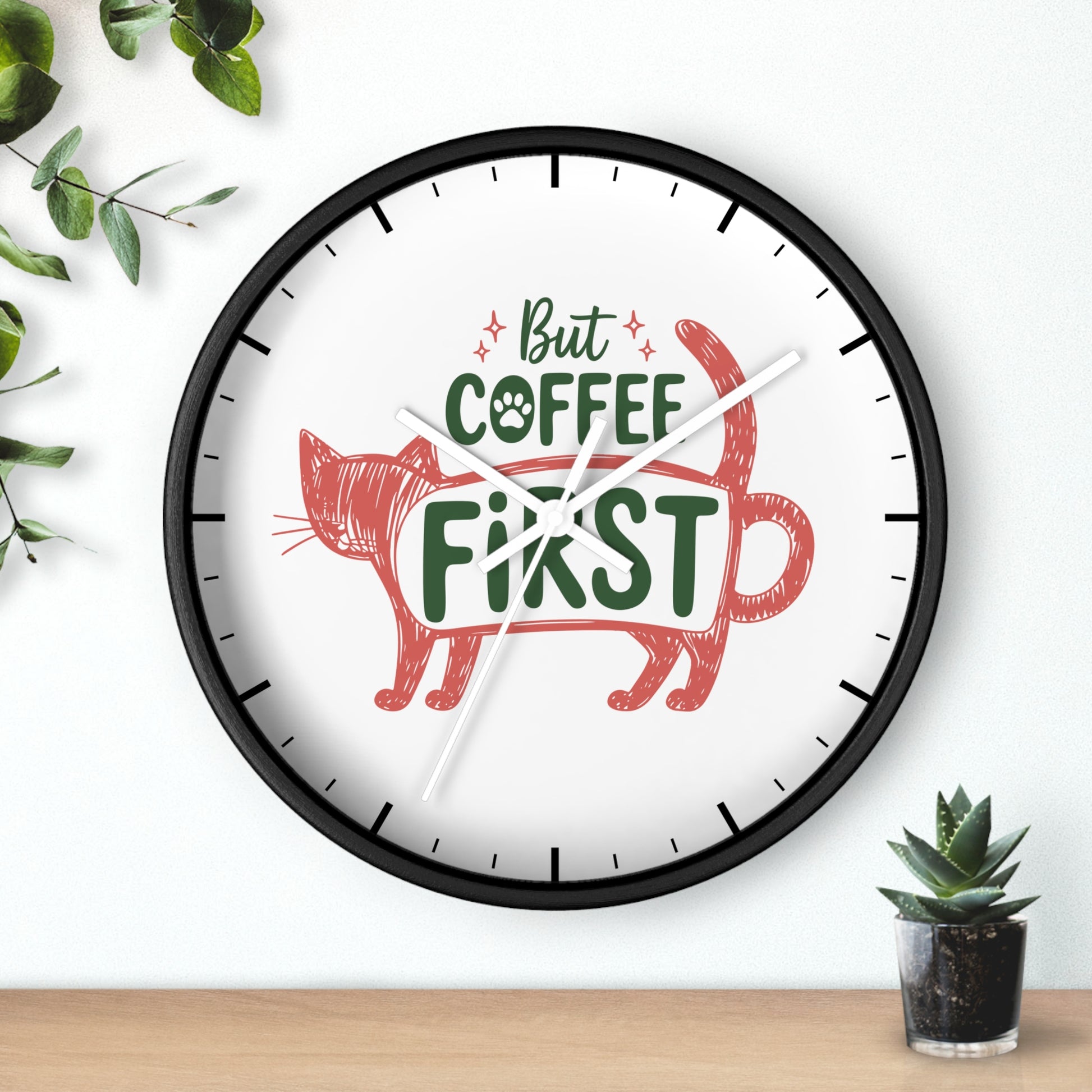 A round wall clock with a black frame and a white face, featuring the phrase 'But COFFEE FIRST' in green and red lettering with a red cat illustration. The clock is mounted on a white wall above a light wood surface, with decorative green leaves and a small potted succulent nearby.