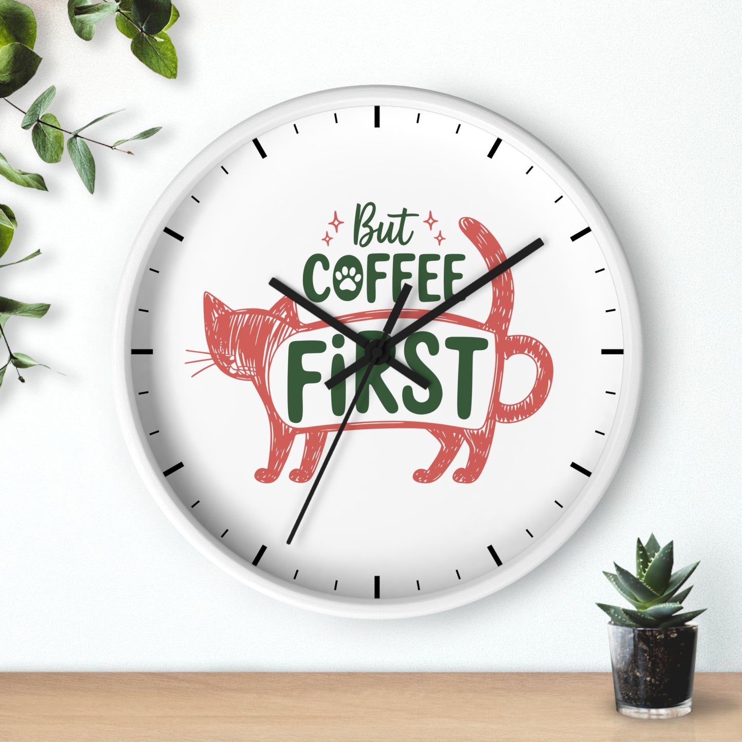 A round white wall clock with black hands and black hour markers, featuring the phrase 'But COFFEE FIRST' in green and red lettering above a red cat illustration. The clock is shown hanging on a white wall above a light wood surface, with green leaves and a small potted succulent as decorative props.