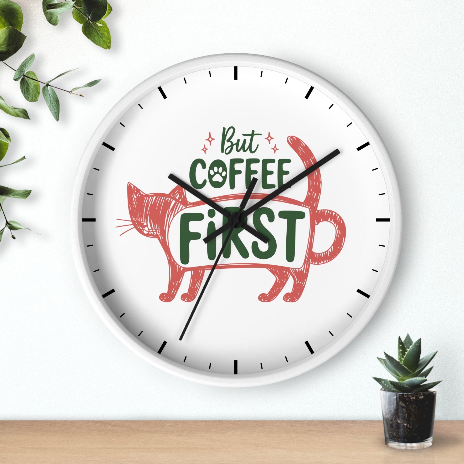 A round white wall clock with black hands and black hour markers, featuring the phrase 'But COFFEE FIRST' in green and red lettering above a red cat illustration. The clock is shown hanging on a white wall above a light wood surface, with green leaves and a small potted succulent as decorative props.
