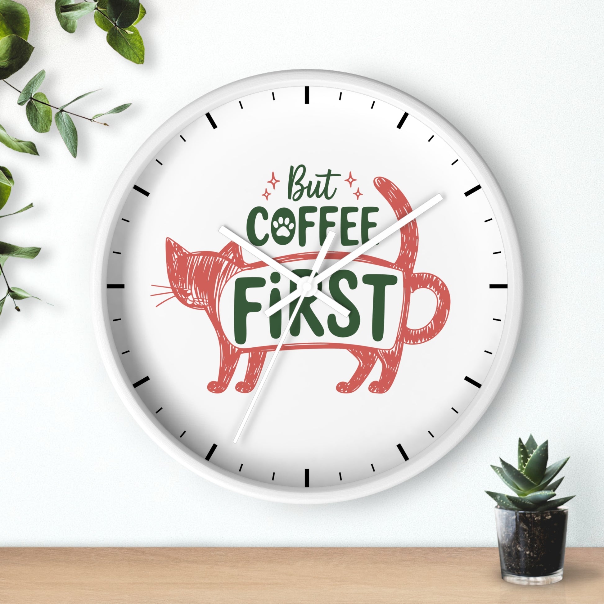 A round white wall clock with black hands and black hour markers, featuring a red sketch-style cat silhouette and the phrase 'But COFFEE FIRST' in green and red text. The clock is shown hanging on a white wall above a light wood surface, with green leaves and a small potted succulent as props.