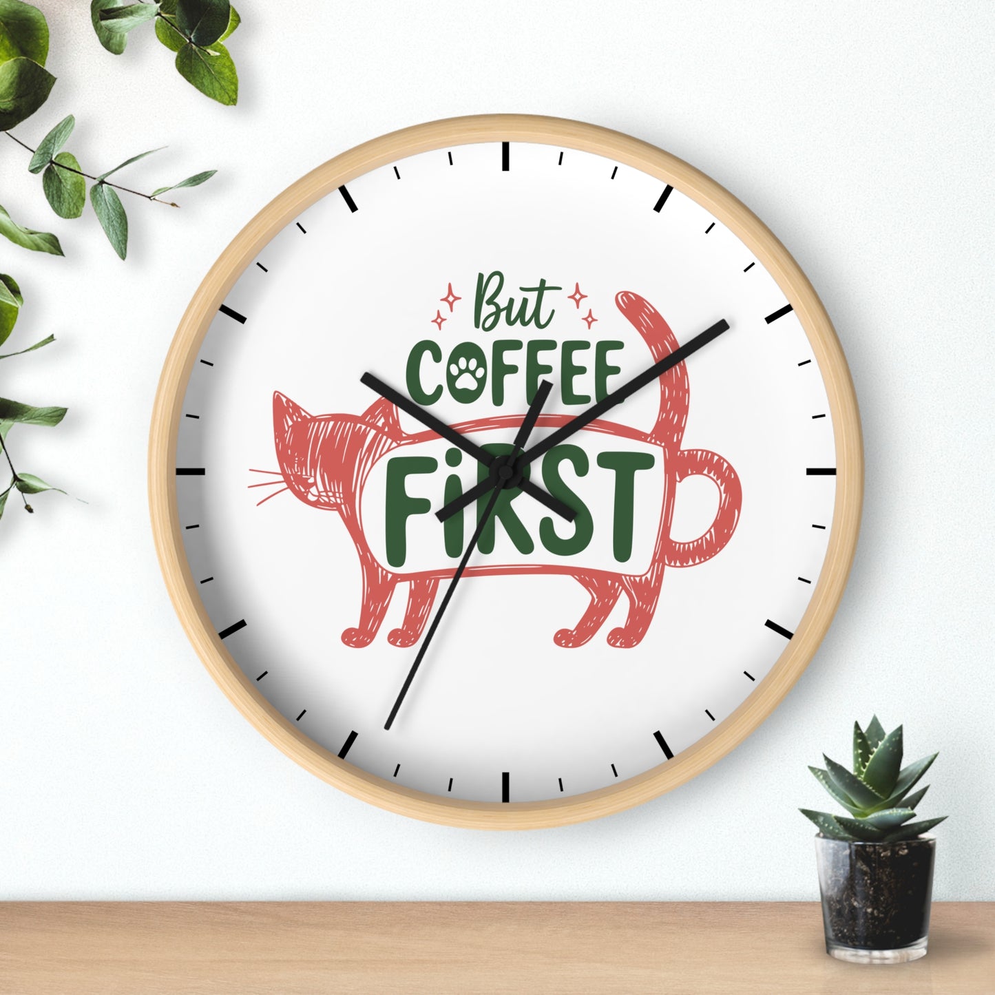 A round wall clock with a light wood frame and a white face, featuring the phrase 'But COFFEE FIRST' in green with a red cat illustration. The clock is mounted on a white wall above a wooden surface, with decorative green leaves and a small potted succulent nearby.