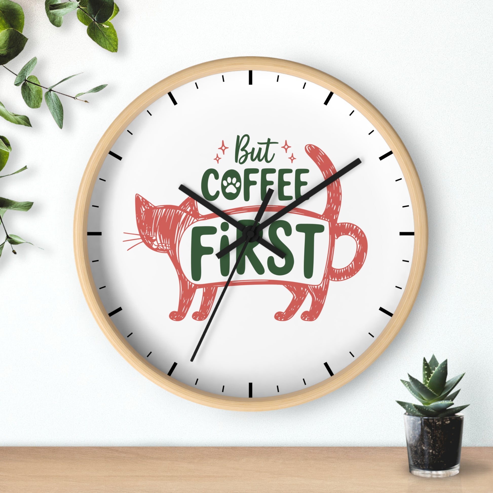 A round wall clock with a light wood frame and a white face, featuring the phrase 'But COFFEE FIRST' in green with a red cat illustration. The clock is mounted on a white wall above a wooden surface, with decorative green leaves and a small potted succulent nearby.